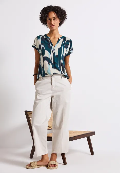 Frau mit gemustertem Shirt, heller Hose und Sandalen steht vor einem Stuhl.